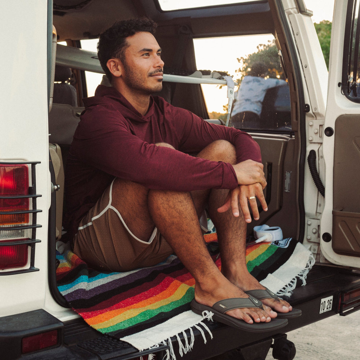 Man in maroon hoodie and brown shorts sitting in open van door wearing brown koa sandals.