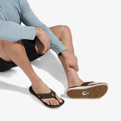 Man wearing light blue long-sleeve shirt and black shorts seated, displaying black flip-flop sandal with tan sole.