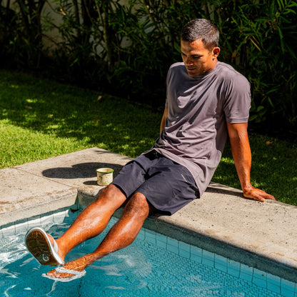 Man in ash gray t-shirt and dark shorts sitting poolside with feet in water.
