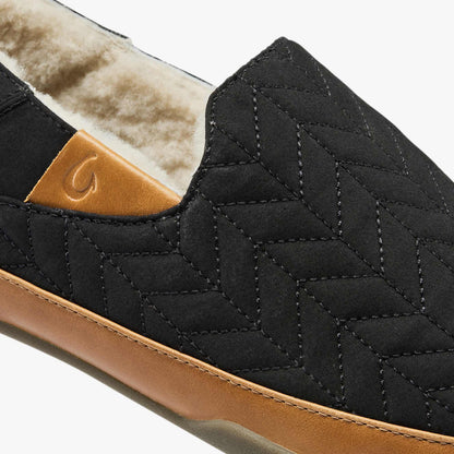 Black quilted slipper with tan leather sole and cream fleece lining, showing curved stitching detail and logo patch.