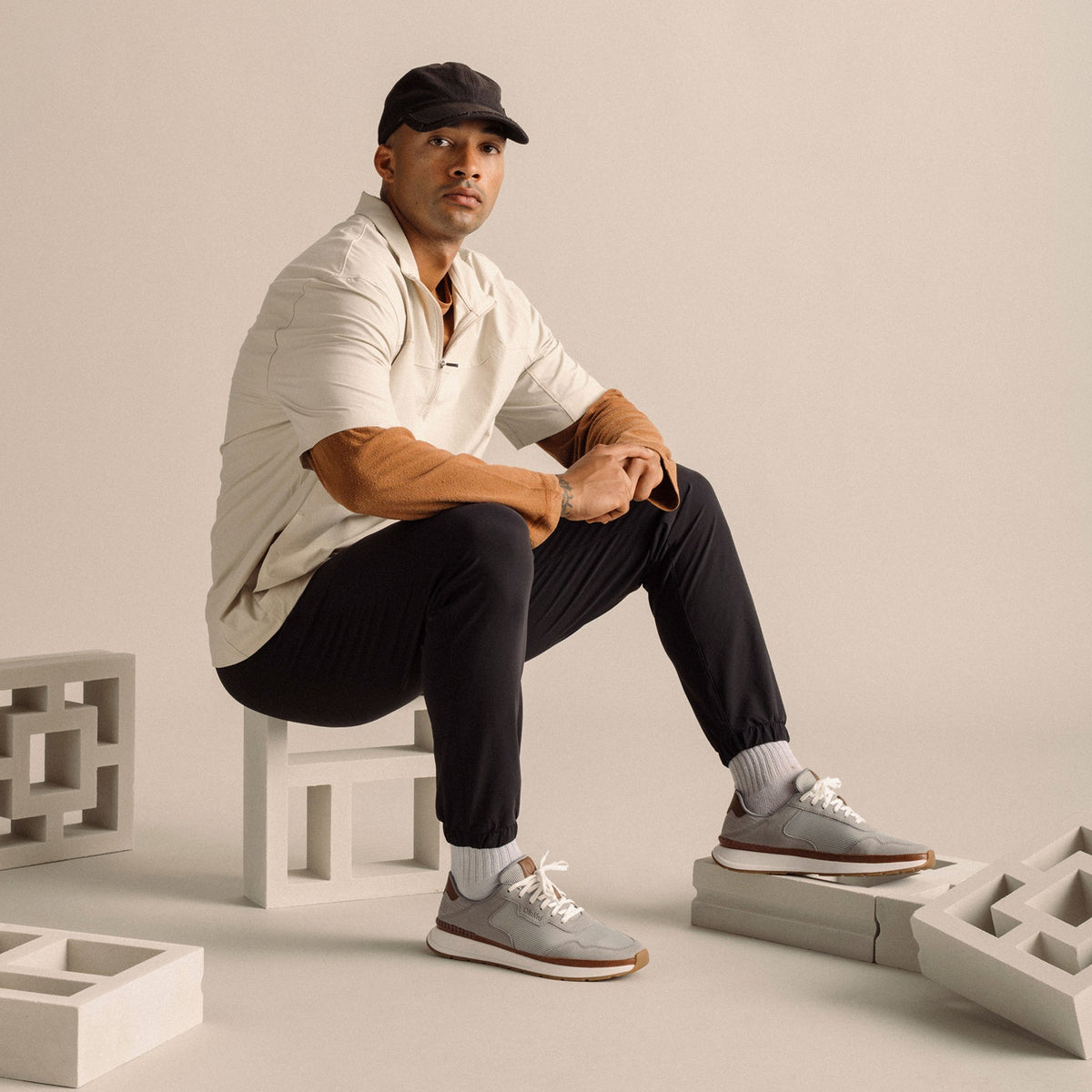 Man wearing light grey sneaker with brown trim, black pants, white shirt, and black cap, sitting on white block.