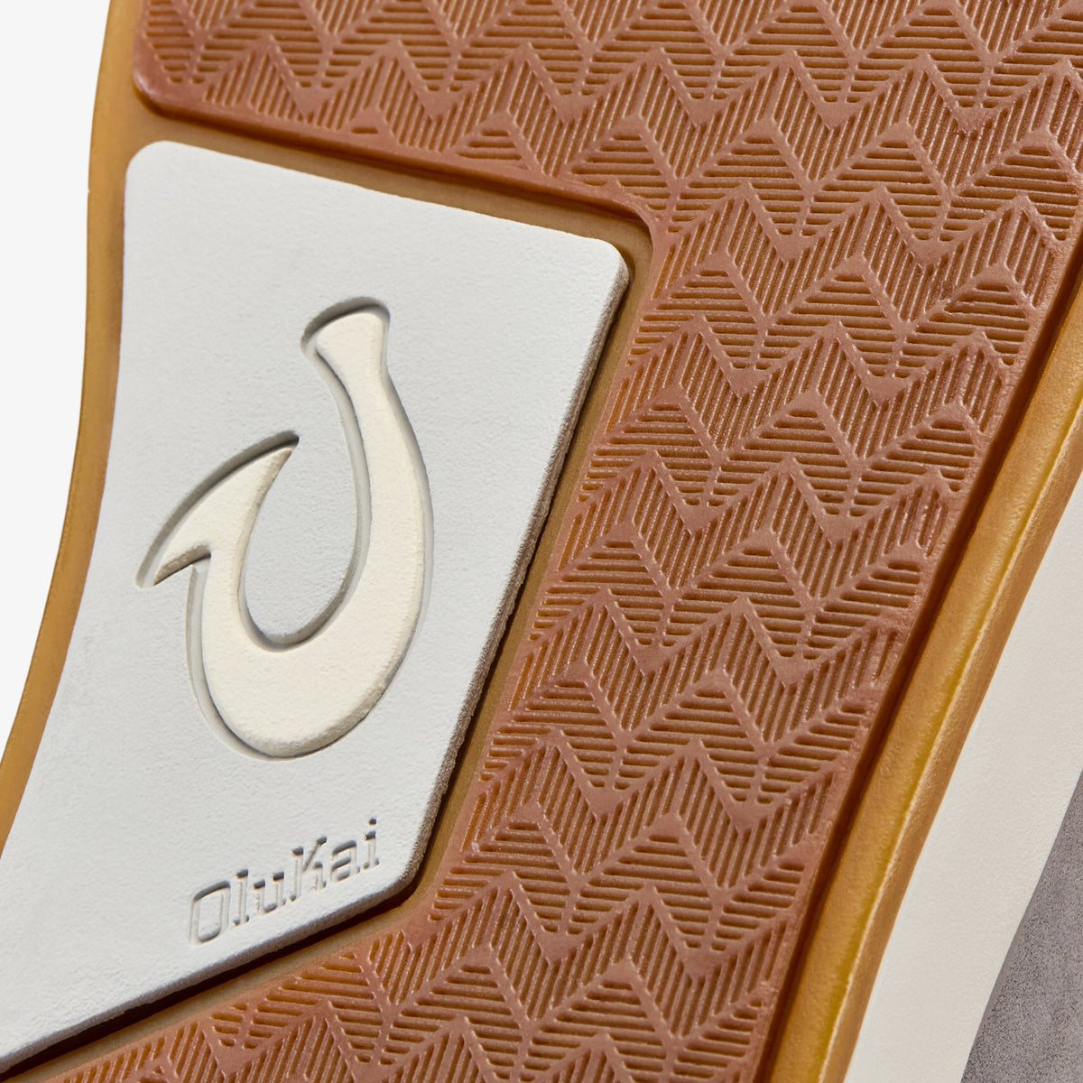 Tan rubber sole with geometric diamond pattern and white leather panel featuring embossed hook design and "oluKai" branding.